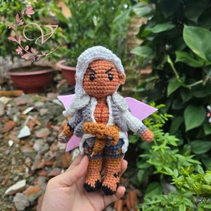 Può includere: Bambola all'uncinetto fatta a mano raffigurante un personaggio con cappuccio bianco, giacca grigia e bianca e pelle arancione. La bambola ha un'ala rosa e indossa pantaloncini blu e stivali marroni. La bambola è tenuta in mano.