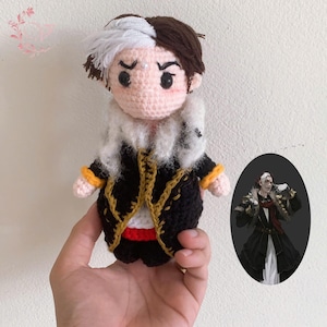 May include: Crocheted doll with white and brown hair, wearing a black and gold coat with a white fur collar. The doll is holding a yellow and gold accessory in its right hand. The doll is standing on a hand.