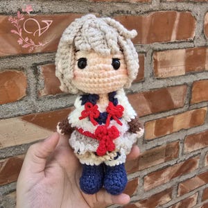 May include: A handmade crochet doll with a light beige face and hair, black eyes, and a white and blue outfit with red accents. The doll is wearing blue boots and brown sleeves. The doll is approximately 5 inches tall.