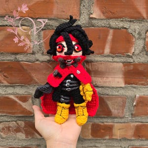 May include: A crocheted plush toy of a character with black hair, a red cape, and yellow boots. The character has a black mask with red eyes and a red bow tie.