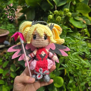 Può includere: Bambola all'uncinetto fatta a mano con capelli biondi, occhi blu e un vestito rosso e rosa. La bambola ha ali rosa e grigie e tiene in mano un bastone rosa. La bambola è un personaggio di un videogioco.