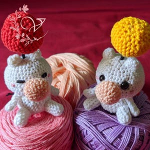 May include: Two crocheted white stuffed animals with pink noses and black eyes. One animal has a red crocheted ball on its head and is sitting on a pink yarn ball. The other animal has a yellow crocheted ball on its head and is sitting on a purple yarn ball.
