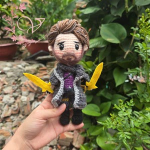 Op de afbeelding: Handgemaakte gehaakte pop van een personage met bruin haar en een baard, gekleed in een grijze jas met een paars shirt en een zwarte broek. De pop houdt twee gele zwaarden vast. De pop is ongeveer 15 cm hoog.