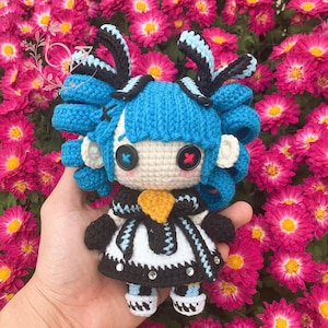 Może przedstawiać: Lalka szydełkowa z niebieskimi włosami, czarno-białą odzieżą i żółtą kokardką. Lalka ma duże oczy z czarnymi i czerwonymi źrenicami. Lalka siedzi w dłoni na tle różowych kwiatów.