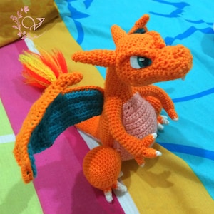 Könnte beinhalten: Orangefarbenes und türkisfarbenes gehäkeltes Charizard Pokemon Plüschtier mit einem Flammenschwanz.