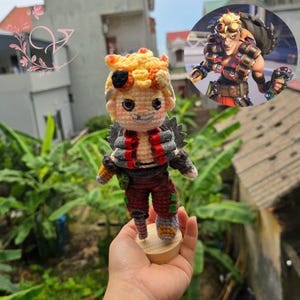 May include: A handmade crochet doll of a character with blonde hair, a black eyepatch, and a mischievous grin. The doll wears a red and gray outfit with a detailed design, standing on a small wooden base. A reference image of the character is in the background.
