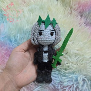 May include: Crocheted doll with gray hair, a green crown, and a green sword. The doll wears a black jacket, black pants, and gray boots. The doll is handmade and has a cute, stylized design.
