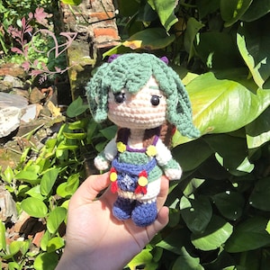 May include: A handmade crochet doll with green hair and a detailed outfit. The doll has a light skin tone, with a green and blue outfit, and a small bag. The doll is held in front of a background of green leaves.