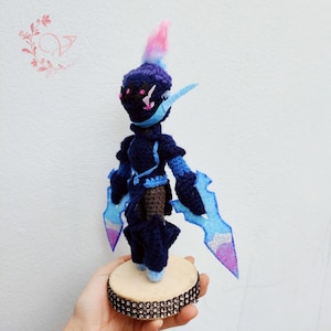 May include: A handmade crochet doll in shades of navy blue, light blue, and pink. The doll has a unique design with a spiky head, large blue blades, and is mounted on a wooden base with a rhinestone border. The doll is approximately 6 inches tall.
