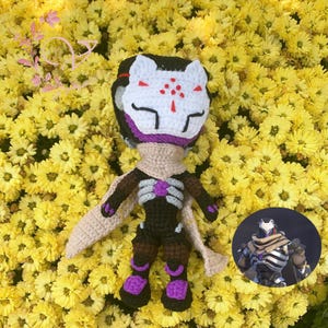May include: A handmade crochet doll with a white and red fox mask, black hair, and a beige scarf. The doll has a black and brown outfit with purple accents. The doll is set against a background of yellow flowers.