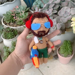Peut inclure: Poupée amigurumi en crochet avec des cheveux bruns, des écouteurs rouges, des lunettes de soleil bleues, une serviette blanche et un pistolet à eau bleu et orange. La poupée porte un short de bain vert et a une barbe brune.