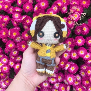 May include: A crocheted doll with brown hair, wearing a yellow jacket, brown pants, and white boots. The doll is holding its hands out and has a friendly expression. The doll is sitting on a hand against a background of pink flowers.