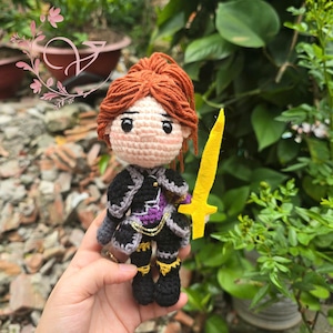 May include: Crocheted doll of a warrior with red hair, wearing black and purple armor, and holding a yellow sword. The doll has a detailed design with gold accents and is set against a green background.