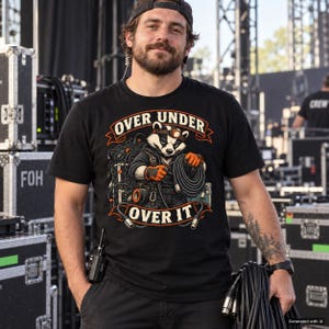 Puede incluir: Una camiseta negra de manga corta con un gráfico de un tejón con linterna frontal y guantes de trabajo, sosteniendo un cable negro enrollado. El texto "OVER UNDER" aparece sobre el tejón y "OVER IT" debajo en letras naranjas y blancas.