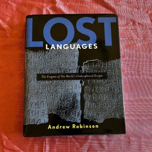 May include: A hardcover book titled "Lost Languages" with blue text on a black background. The cover features an image of ancient inscriptions and the author's name, Andrew Robinson. The book explores the enigma of undeciphered scripts.