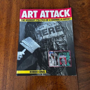 May include: A book titled "ART ATTACK" with the subtitle "THE MIDNIGHT POLITICS OF A GUERRILLA ARTIST." The cover features a black and white image of a person's face with the word "HERE" overlaid. A smaller image says "IT CAN'T HAPPEN HERE."
