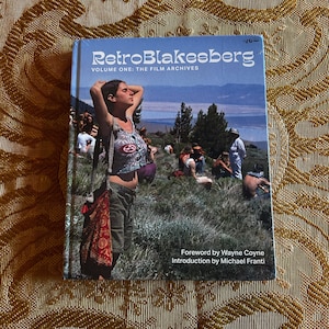 May include: A book titled "RetroBlakesberg Volume One: The Film Archives" with a photograph of a group of people on a hillside. The cover features a woman with her arms raised. The book's foreword is by Wayne Coyne and the introduction by Michael Franti.