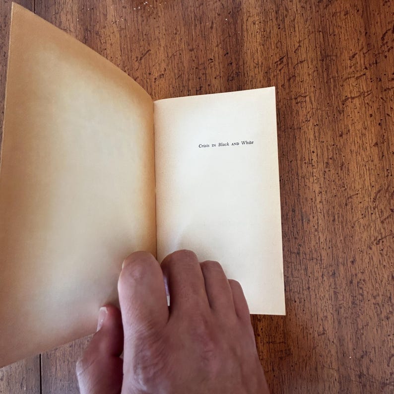 May include: An open book with cream-colored pages featuring the text Crisis in Black and White printed in a simple black serif font on the right-hand page. The paper appears aged with a slight yellow tint, and the book is held open by a hand.