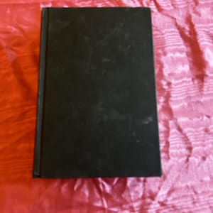Puede incluir: Un libro negro cerrado con una cubierta ligeramente desgastada. El libro es rectangular y parece ser de tapa dura. El fondo es una tela rosa texturizada.