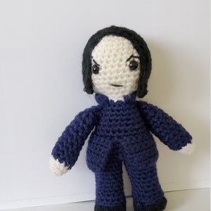 May include: Crocheted doll with black hair, a white face, and a blue suit. The doll has a serious expression.