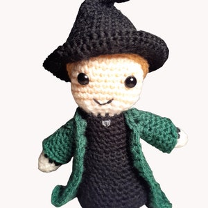 May include: A crocheted plush toy of a young wizard wearing a black pointed hat and a green robe. The toy has a light brown complexion, black eyes, and a small smile.