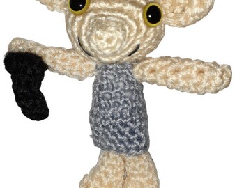 May include: A crocheted plush toy in the shape of a small, humanoid creature with large ears, a gray shirt, and yellow eyes. The toy is holding a black object in its left hand.