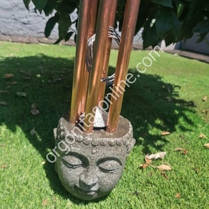 Peut inclure: Un carillon en cuivre à cinq tubes suspendu à une sculpture de tête de Bouddha en pierre. Le carillon est suspendu au sommet de la tête du Bouddha. La sculpture est posée sur une surface herbeuse verte. Le texte "orgoniteonline.com" est visible sur l'image.
