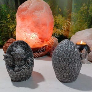 Puede incluir: Dos portavelas de huevo de dragón grises con una superficie texturizada. Los portavelas están sobre una superficie blanca. Una lámpara de sal del Himalaya está en el fondo.
