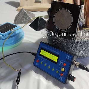 Könnte beinhalten: Ein blauer FG-200 DDS Funktionsgenerator mit Digitalanzeige, die "Mavet Schumann Freat 07.83Hz" anzeigt. Ein schwarzes Quadrat mit Spiralmuster liegt auf einem Stein. Ein blaues und weißes Gerät mit Solarpanel ist ebenfalls vorhanden.