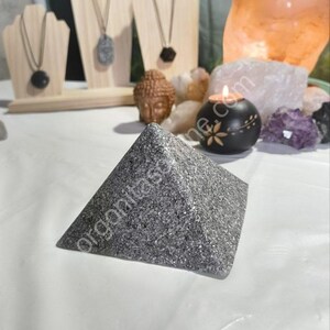 May include: A gray pyramid-shaped crystal with a sparkly, textured surface. The crystal is sitting on a white surface.