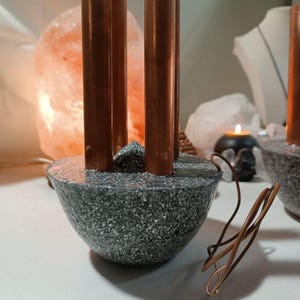 Puede incluir: Un objeto decorativo con cuatro tubos de cobre verticales incrustados en una base gris moteada en forma de cuenco. Una pequeña piedra gris puntiaguda se sitúa en el centro entre los tubos y un alambre de cobre enrollado descansa cerca de la base.