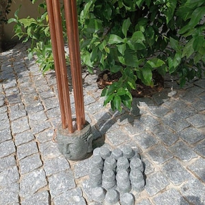 Op de afbeelding: Een windgong met drie lange, verticale koperen buizen in een grijze stenen voet in de vorm van een Boeddha-hoofd, vergezeld door een verzameling van zestien kleine, ronde, grijze stenen decoratieve stukken die ernaast zijn geplaatst.