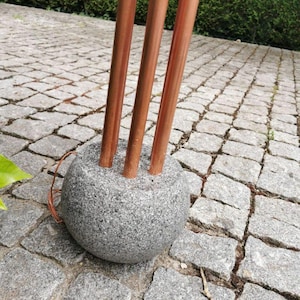 Op de afbeelding: Een bolvormige grijze granieten stenen voet met een getextureerd, gespikkeld oppervlak. Drie verticale, gladde, gepolijste koperen buizen zijn in de bovenkant van de steen verwerkt. Een dunne koperdraad steekt uit de zijkant van de stenen voet.