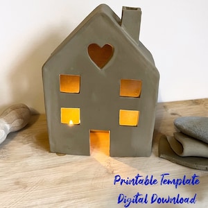 May include: Printable template for a house-shaped candle holder. The template is for a digital download. The image shows a grey clay house with a heart-shaped cutout and windows. The house is lit from inside with a candle.