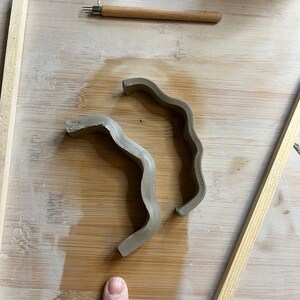 May include: Two pieces of brown clay shaped into wavy, curved forms on a wooden surface.