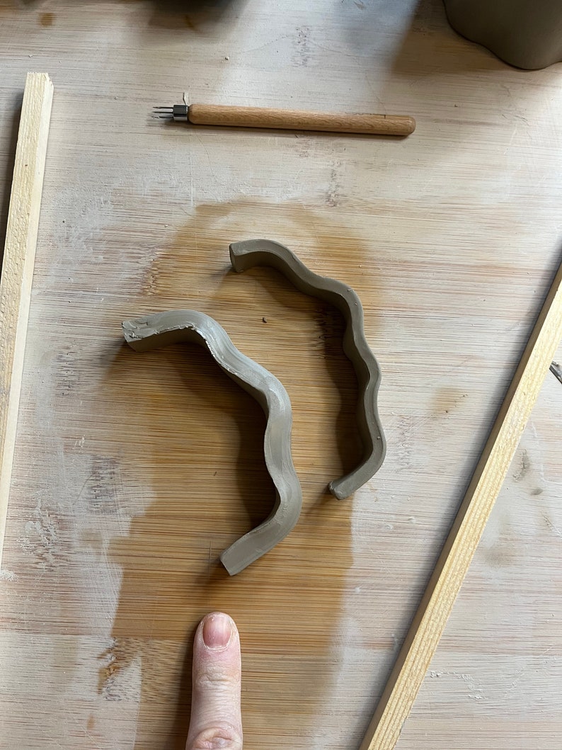 May include: Two pieces of brown clay shaped into wavy, curved forms on a wooden surface.