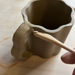 May include: A brown clay mug with a handle being smoothed with a wooden tool. The mug has a wavy rim and a textured handle.