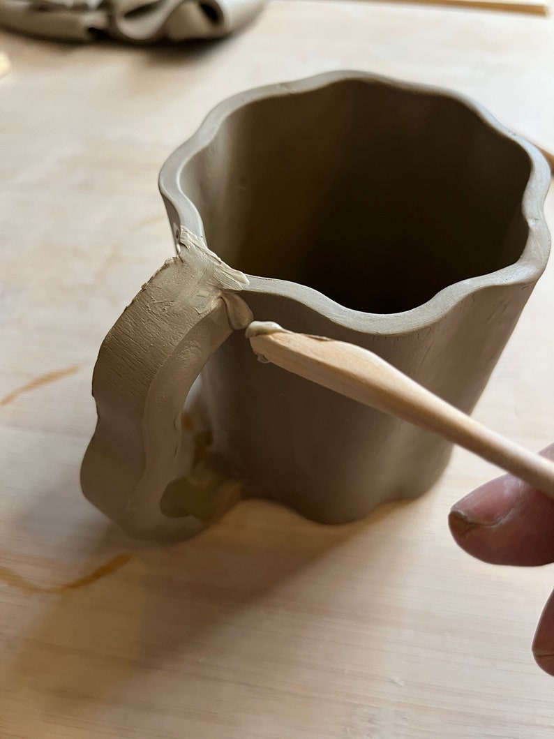 May include: A brown clay mug with a handle being smoothed with a wooden tool. The mug has a wavy rim and a textured handle.