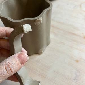 May include: A brown ceramic mug with a handle being attached to the mug. The handle is made of clay and has been attached with a white slip.