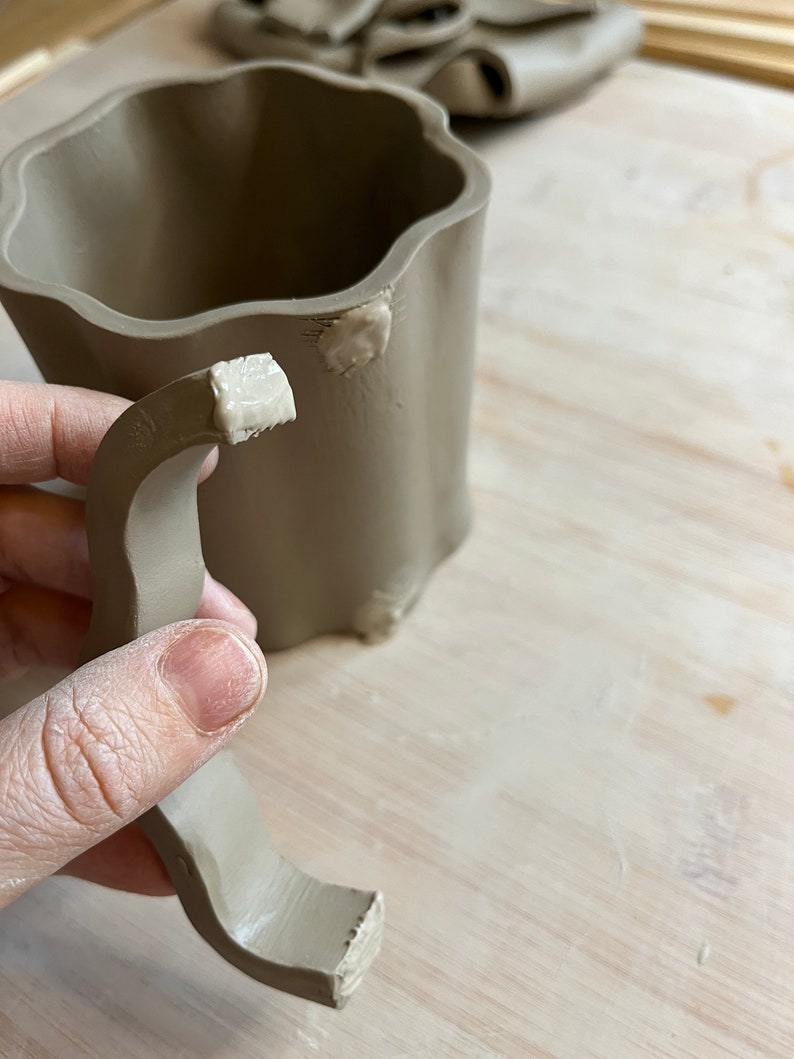 May include: A brown ceramic mug with a handle being attached to the mug. The handle is made of clay and has been attached with a white slip.