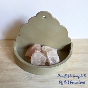 May include: A light grey concrete wall shelf with a scalloped top and a rounded bottom. The shelf is designed to be hung on a wall and has a small hole at the top for hanging. The shelf is filled with four pink rose quartz crystals.