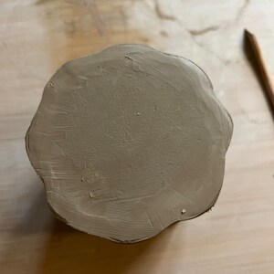 May include: A round, brown clay pot with a scalloped edge. The pot is sitting on a light brown surface.