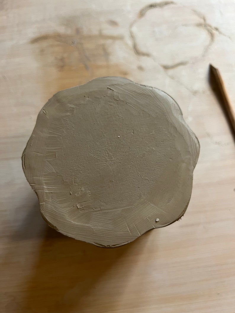 May include: A round, brown clay pot with a scalloped edge. The pot is sitting on a light brown surface.