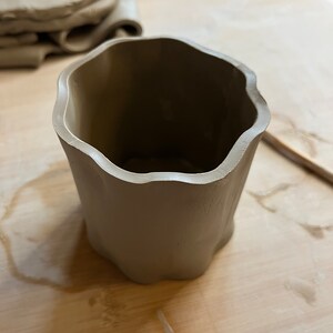 May include: A light brown ceramic cup with a wavy rim. The cup is sitting on a wooden surface.