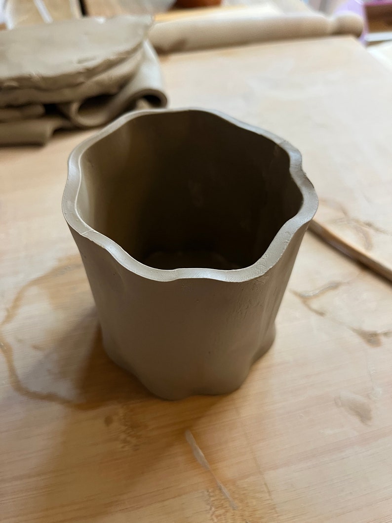 May include: A light brown ceramic cup with a wavy rim. The cup is sitting on a wooden surface.