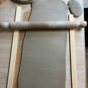 May include: A brown clay slab is being rolled out on a wooden surface using a wooden rolling pin. Two wooden strips are placed on either side of the clay to help guide the rolling pin. A small brown rock is also visible in the image.
