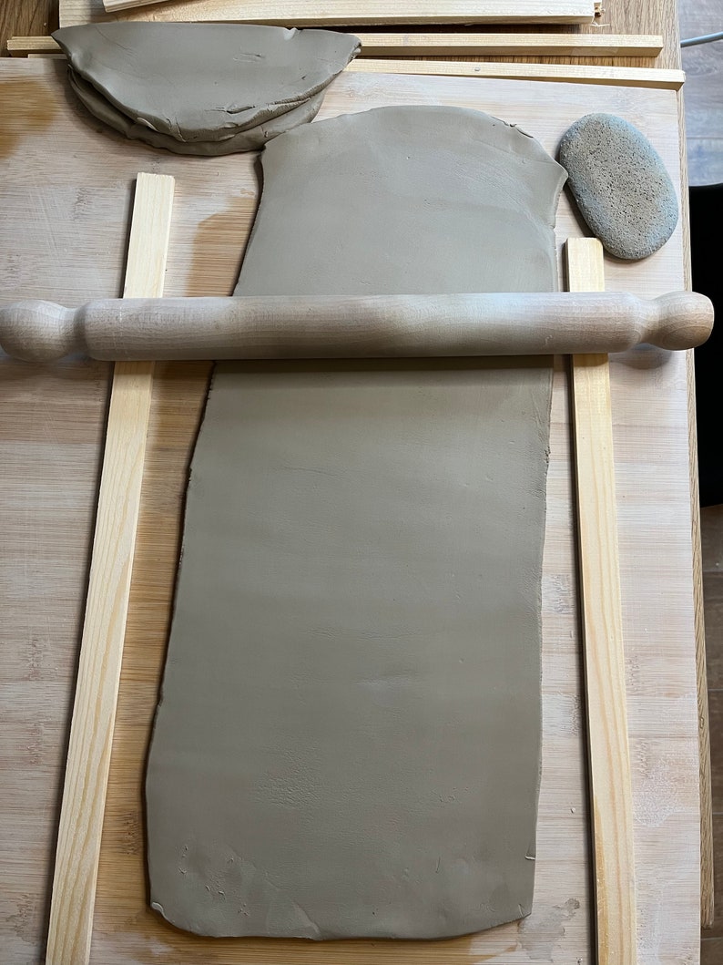May include: A brown clay slab is being rolled out on a wooden surface using a wooden rolling pin. Two wooden strips are placed on either side of the clay to help guide the rolling pin. A small brown rock is also visible in the image.