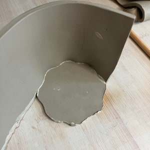 May include: A gray ceramic bowl with a rounded edge is being attached to a flat piece of clay. The clay is on a light wood surface.