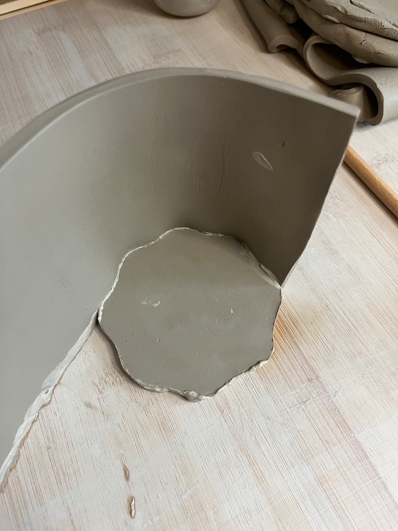 May include: A gray ceramic bowl with a rounded edge is being attached to a flat piece of clay. The clay is on a light wood surface.