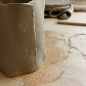May include: A close-up of a light brown ceramic pot with a textured surface. The pot is sitting on a wooden table.